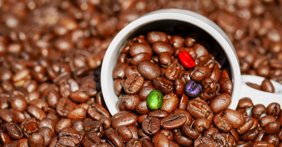 A lot of coffee beans with a partially filled cup laying on it. 3 beans in a cup have different color